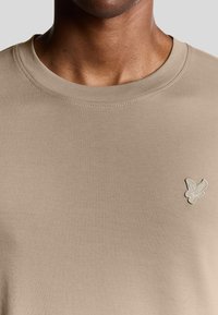 Khakifarbenes Baumwoll-T-Shirt mit Rundhalsausschnitt, das ein kleines gesticktes Vogel-Logo auf der linken Brust zeigt. Glatte Textur und einfaches Design.