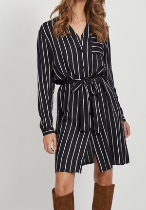 Femme portant une robe chemise noire à rayures verticales blanches, avec une ceinture à la taille et des bottes marron à hauteur du genou.