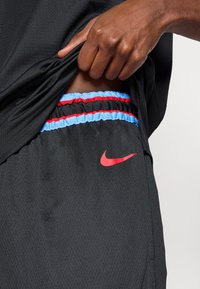 Juodi sportiniai šortai su raudonu Nike logotipu, su mėlynu ir raudonu elastingu dirželiu. Atrodo, kad medžiaga yra lengva ir tekstūruota.