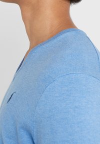 Ljusblå V-ringad t-shirt i mjukt tyg, med en liten mörk logotyp på vänster bröst och en ribbad textur vid halsringningen.