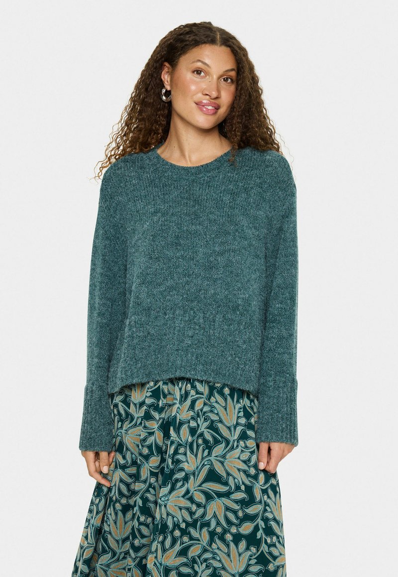 Türkisfarbener gestrickter Pullover mit rundem Halsausschnitt und langen Ärmeln, der eine strukturierte Oberfläche aufweist; kombiniert mit einem Rock im Blumenmuster.