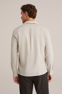 Camicia a maniche lunghe color beige chiaro con colletto, piega sul retro e polsini abbottonati, realizzata in tessuto liscio e morbido. Abbinata a pantaloni scuri.
