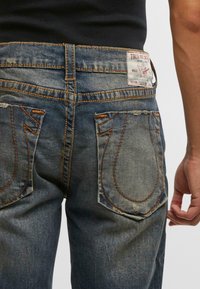 Pohľad zozadu na osobu, ktorá má na sebe vyblednuté modré džínsy s výrazným oranžovým prešitím a bielou značkovou etiketou True Religion na páse.