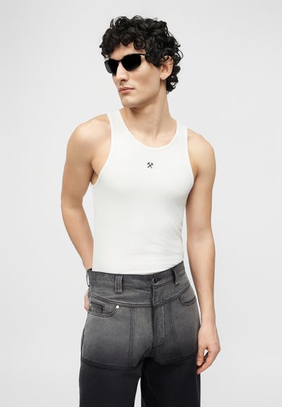 Άνδρας με σγουρά μαλλιά, που φοράει μαύρα γυαλιά ηλίου, λευκό αμάνικο πουκάμισο με μικρό λογότυπο και σκουρόχρωμα τζιν με διαβαθμισμένη απόχρωση, όρθιος μπροστά σε απλό φόντο.