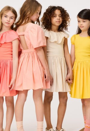 Cuatro niñas llevan vestidos coloridos: un vestido melocotón con volantes, un vestido beige con falda acampanada, un vestido amarillo y calcetas rosas claras.