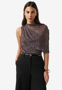 Brown polka dot blouse with draped design, sheer texture, and one-shoulder style, paired with high-waisted black trousers and a gold bracelet.