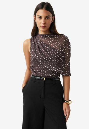 Brown polka dot blouse with draped design, sheer texture, and one-shoulder style, paired with high-waisted black trousers and a gold bracelet.