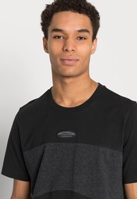 Fekete és szürke adidas póló, kerek nyakkal, rövid ujjal, textúrált anyagból készült, középen a logóval ellátott emblémával.