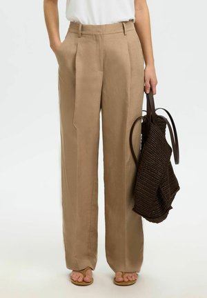 Kvinde iført beige højtaljede vidde bukser og hvid top, holder en mørkebrun vævet taske og står barfodet på hvid baggrund.