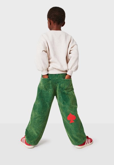 Bambino di spalle con le mani in tasca, che indossa pantaloni in corda verde a gamba larga con simbolo a forma di picche rossa e sneakers rosse con strisce bianche.