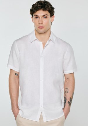 Młody mężczyzna z kręconymi włosami, ubrany w białą koszulę na krótki rękaw z guzikami i beżowe spodnie, stojący z rękami w kieszeniach.