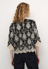 Svart blus med intrikata vita spetsbroderier med paisleymönster, vågformad spetskant och trekvartsärmar.