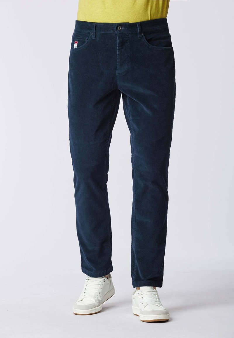 Pantaloni in corduroy blu scuro con design slim fit, dotati di tasche laterali e chiusura con bottone. Indossati con sneakers bianche.