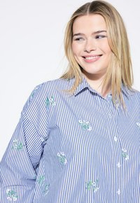 Camicia button-up a righe blu e bianche con ricami floreali verdi e bianchi, dotata di colletto e maniche lunghe.