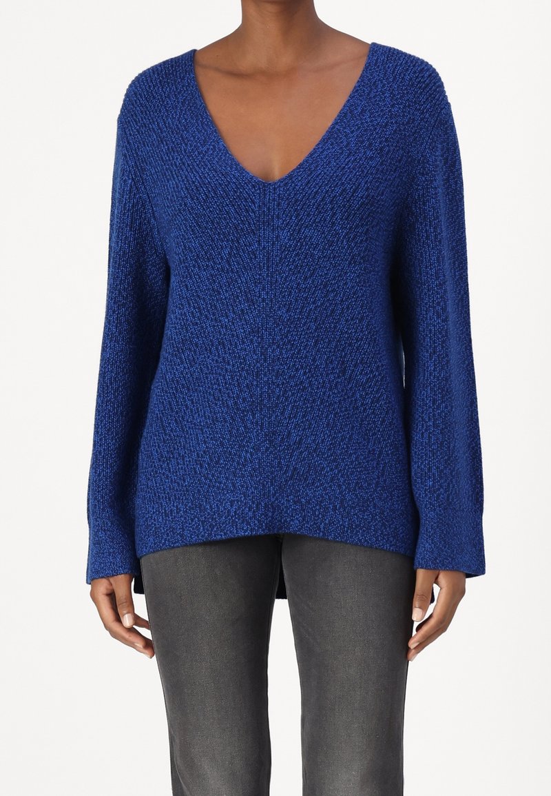 Femme portant un pull à manches longues texturé bleu royal avec un col en V, associé à un pantalon gris foncé, debout devant un fond uni.