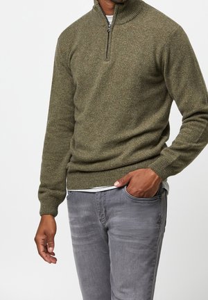 Homme portant un pull vert olive à fermeture éclair partielle et un jean gris, une main dans la poche, sur un fond blanc uni.