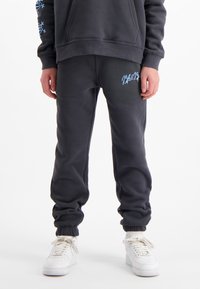 Donkergrijze sweatpants gemaakt van zacht materiaal, met een elastische tailleband en boorden, voorzien van een blauw geborduurd logo op de dij. Gepaard met witte sneakers.