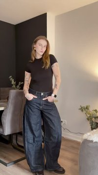 T-shirt preta justa usada com jeans de ganga escura de pernas largas, com cintura alta, bolsos grandes e costura mínima. Fundo em chão de madeira.