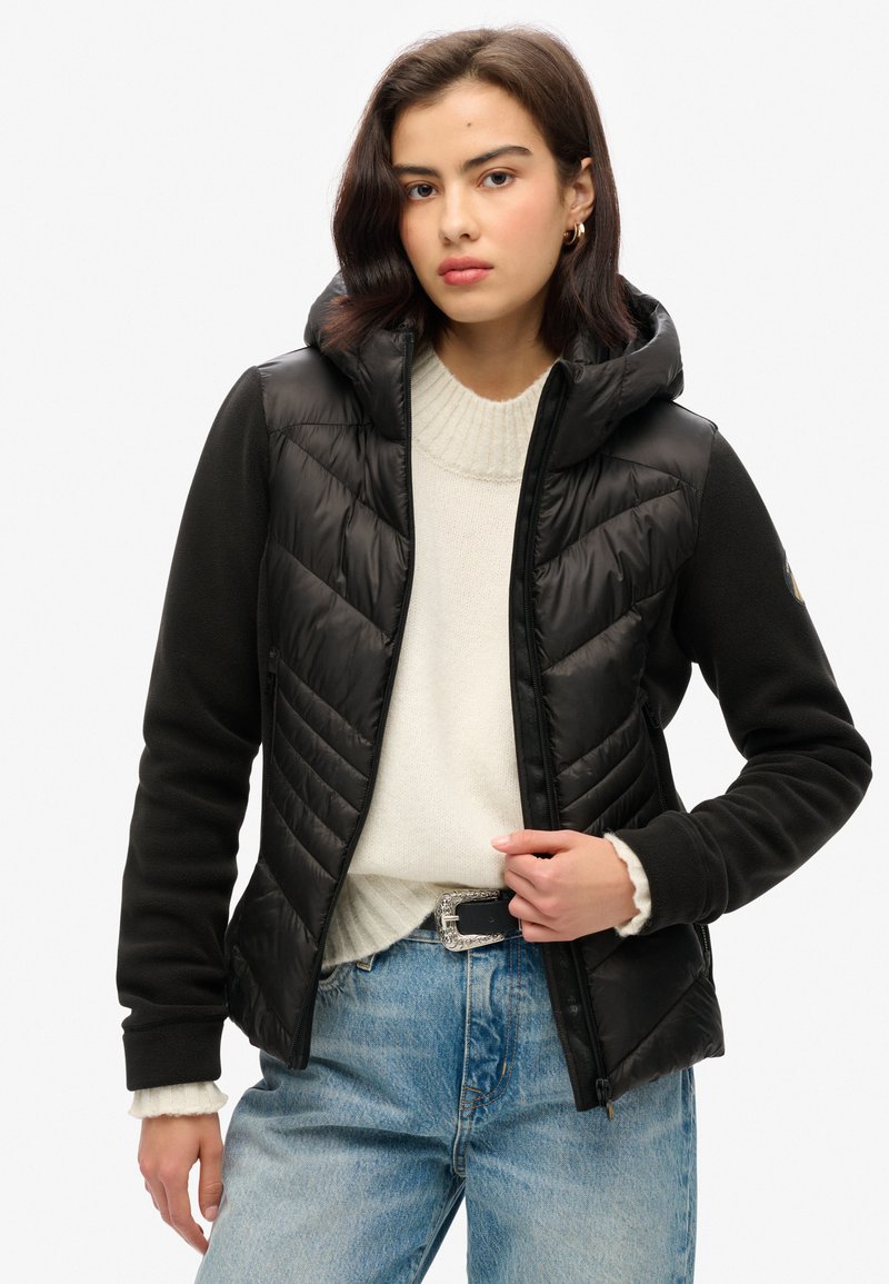Chaqueta de plumas negra con capucha, con diseño de acolchado en forma de chevron, mangas de forro polar y cremallera frontal. Combinada con un suéter crema y jeans claros.