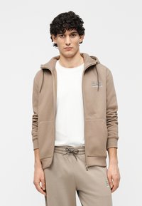 Beige zip-up hoodie met een zachte textuur, voorzien van een capuchon met trekkoord, voorzakken en een klein geborduurd logo op de borst.