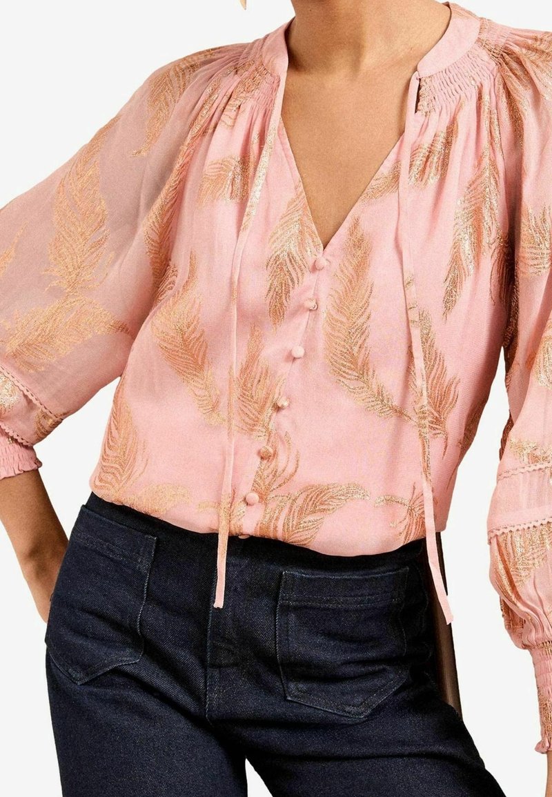 Blouse rose avec motifs de plumes dorées, devant boutonné, col à nouer, manches bouffantes, associée à un jean bleu foncé.