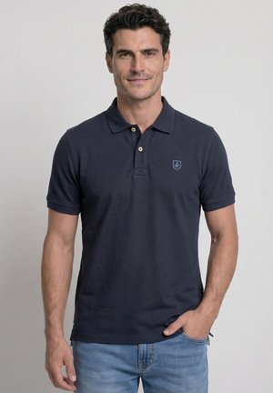 Hombre con cabello rizado oscuro que lleva una camiseta polo azul marino y vaqueros azul claro, de pie con una mano en el bolsillo frente a un fondo liso.