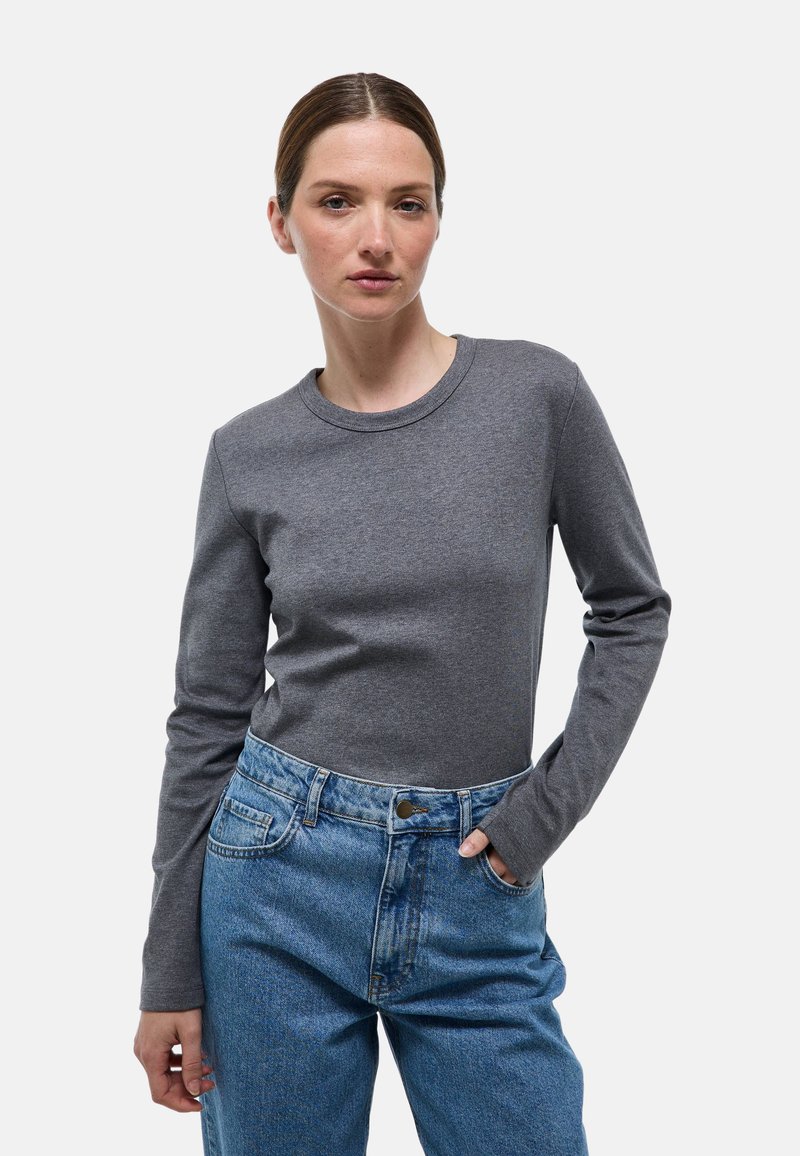 Chemise grise à manches longues avec un col rond, associée à un jean bleu taille haute. Le haut présente une texture lisse et une coupe ajustée.