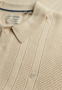 Polo en tricot beige présentant des motifs texturés, un col uni et un logo brodé sur le côté gauche. Boutons au niveau du cou.