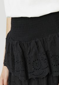 Jupe noire à volants avec une taille smockée, ornée de motifs en broderie anglaise. La texture du tissu est légère et légèrement froissée.