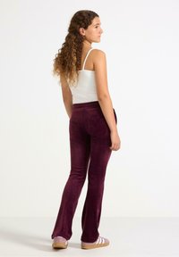 High-waisted, flared velvet pants in deep burgundy, featuring a smooth texture and two back pockets. Paired with a white tank top and casual footwear.