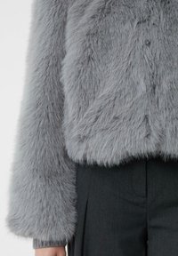 Szara kurtka z sztucznego futra o miękkiej, puszystej teksturze i skrojonym kroju. Posiada szerokie rękawy oraz zaokrąglony dół, zestawiona z ciemnymi spodniami.