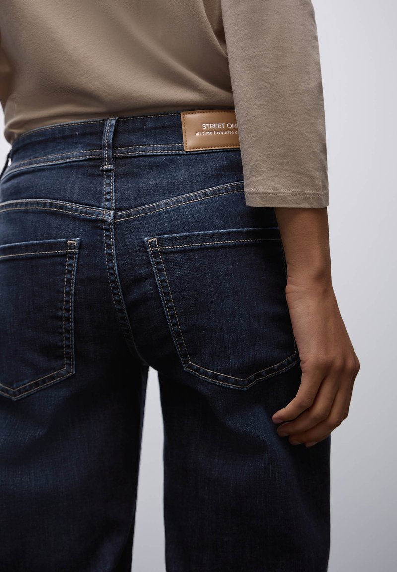 Jeans blu scuro con una texture liscia, dotati di un'etichetta in pelle rettangolare sulla parte posteriore della vita e dettagli di cucitura sottili sulle tasche.