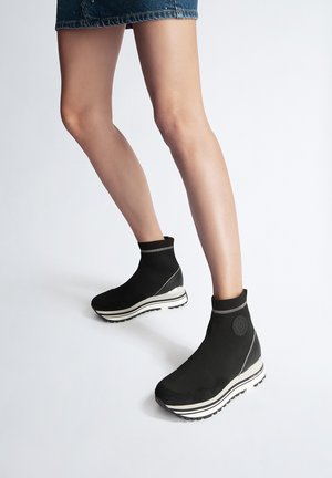 Beine in einem kurzen Jeansrock und schwarzen Plateausneakern im Sockendesign mit weißen, gestreiften Sohlen, stehend auf weißem Hintergrund.