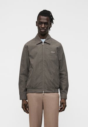 Jeune homme portant une veste zippée marron à carreaux et un pantalon beige, debout devant un fond clair uni.