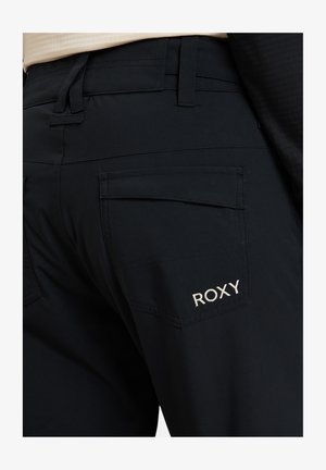 Pantalons noirs avec une texture lisse, une coupe ajustée et de grandes poches arrière. Le logo Roxy est brodé en blanc sur la poche droite.