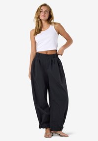 Femme debout, portant un crop top blanc sans manches, un pantalon noir ample taille haute et des sandales à talons ouvertes, sur un fond uni.