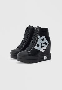Crne wedge tenisice s debelim potplatima, vezanjem sprijeda, bočnim patentnim zatvaračima i velikim srebrnim "DKNY" logotipima sa strane i na potplatima.