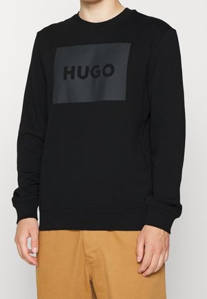 Mężczyzna ubrany w czarną bluzę z długim rękawem z ciemnym prostokątnym logo "HUGO" na piersi, w połączeniu z jasnobrązowymi spodniami.