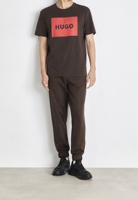 Hnědé tričko s červeným obdélníkovým logem „HUGO“, doplněné o odpovídající hnědé tepláky a černé tenisky s červenými detaily. Jednoduchý design.