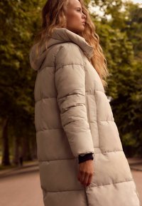 Manteau matelassé beige clair avec capuche, présentant un design matelassé, des coutures visibles et des poches latérales. Le tissu a une texture lisse et douce.
