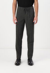 Man in een donkergrijze gestreepte broek met een elastische tailleband en zwarte leren formele schoenen, staande tegen een witte achtergrond.