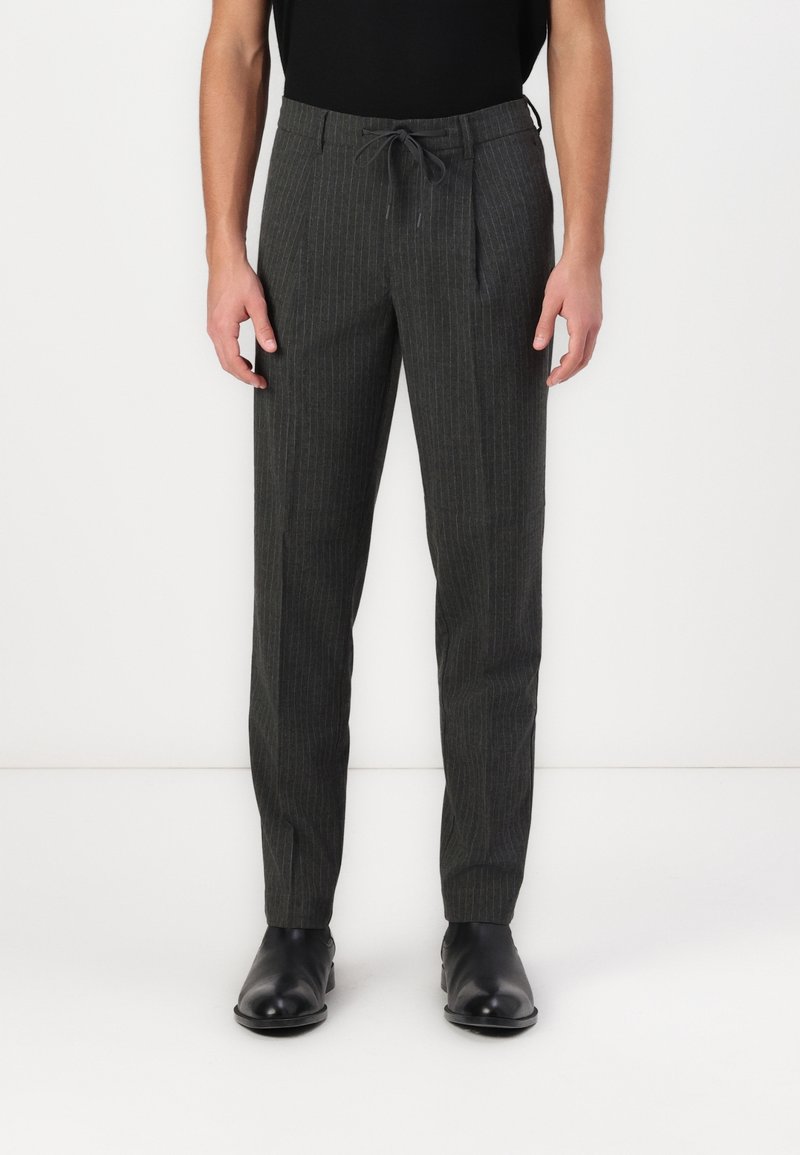 Man in een donkergrijze gestreepte broek met een elastische tailleband en zwarte leren formele schoenen, staande tegen een witte achtergrond.