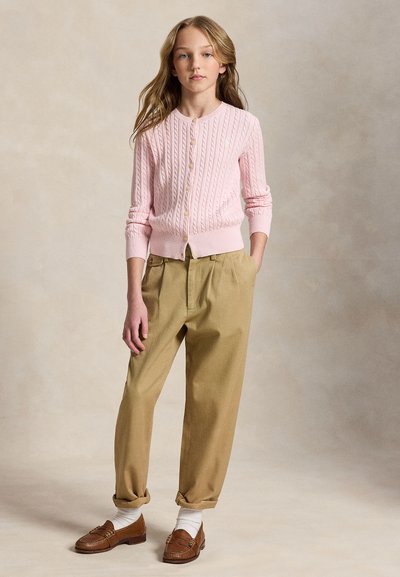 Fiatal lány hosszú hajjal, rózsaszín fonott mintás kardigánt, bézs pliszírozott nadrágot, fehér zoknit és barna mokaszint visel, egyik kezével a zsebében áll.