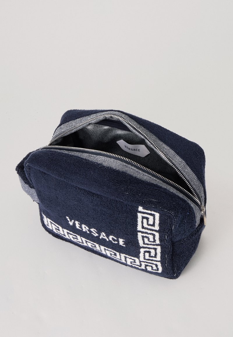 Pochette en tissu bleu marine Versace avec zip frontal, motif clé grecque blanc et étiquette Versace visible à l'intérieur du compartiment principal ouvert.
