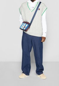 Graue Strickweste mit grünem Akzent über einem weißen Hoodie, kombiniert mit locker sitzenden blauen Denim-Hosen und einer mehrfarbig gestreiften Tasche.