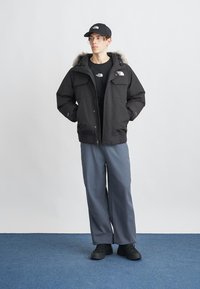 Jachetă neagră izolată cu glugă căptușită cu blană, două buzunare frontale, purtată peste un tricou negru și pantaloni largi de culoare albastru deschis, asortată cu pantofi negri și o șapcă.