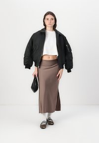 Svart bomberjakke, hvit cropped skjorte, brun midi skjørt, svart veske, hvite sokker og brune leopardmønstrede sko. Glatte teksturer, uformell stil.