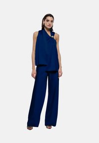 Top senza maniche blu navy con design drappeggiato, abbinato a pantaloni a gamba larga. Include un accento metallizzato sulla spalla. Texture liscia.