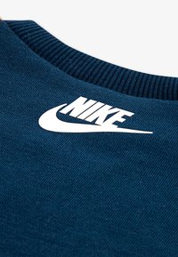 Tamno plavi materijal s teksturiranom površinom. Ima veliki bijeli Nike logo i rebrasti izrez za dodatne detalje.