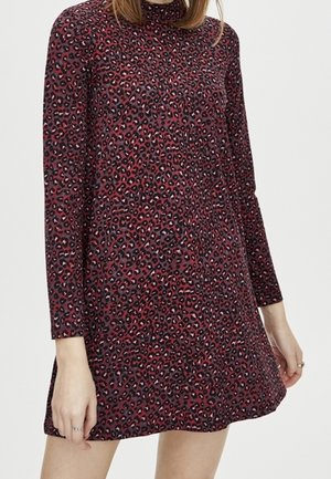 Femme portant une mini-robe à manches longues et col haut avec un motif léopard rouge et noir.